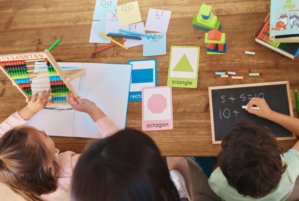 Overhead view of children learning math together with an abacus, geometry cards, worksheets, colorful blocks, and a chalkboard.