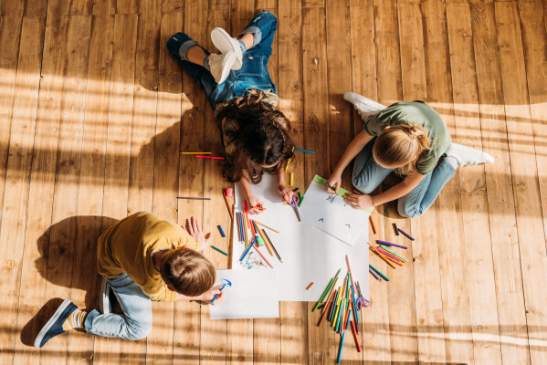 Overhead view of three children drawing together on large paper with colorful pencils on a sunlit wooden floor, creating a playful scene.