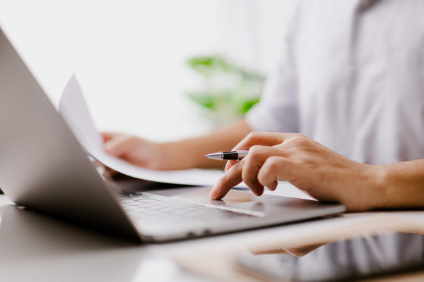 Close-up of a professional using a pen and laptop at a desk, reviewing documents in a bright office; focused, productive atmosphere.