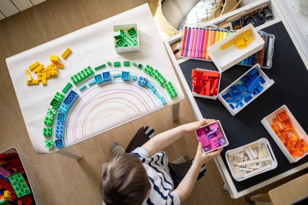 Overhead view of a child sorting colorful building bricks into trays beside a rainbow-patterned design, creating a playful, creative scene.