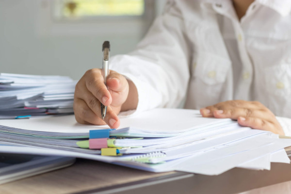 Person in a white shirt writes on paperwork with a pen beside neatly stacked documents and colorful binder tabs, conveying focused office work.